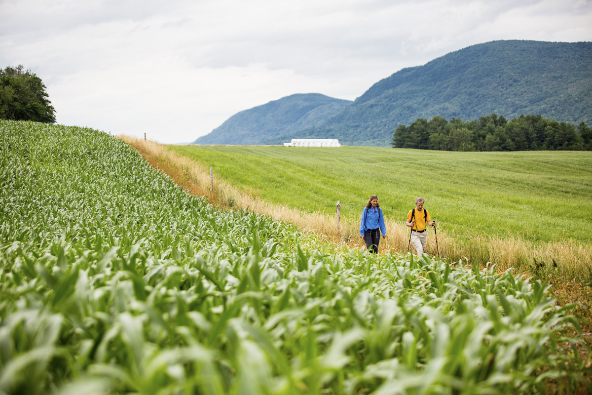 Two people wearing backpacks and hiking gear walk along a path between green fields, with rows of corn on one side and open pasture on the other. Rolling forested mountains rise in the background under a cloudy sky.