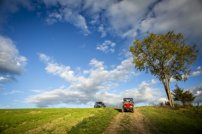 ATV and pickup truck parked on a grassy hill under a wide blue sky, with a tree on the right and clouds scattered overhead.
