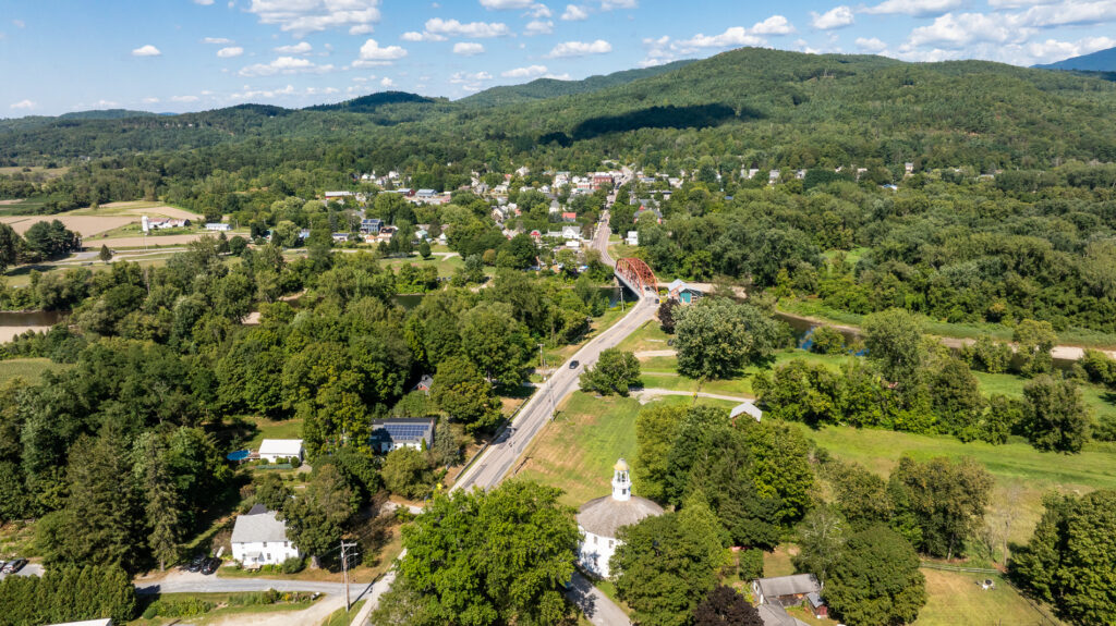 Aerial view of a small Vermont town surrounded by rolling green hills, farmland, and forest. A scenic country road leads into the town, crossing a rust-colored bridge.