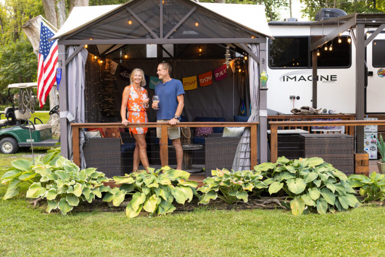 Smiling couple enjoys drinks on a deck outside a cozy RV setup with string lights and flags, surrounded by lush greenery.