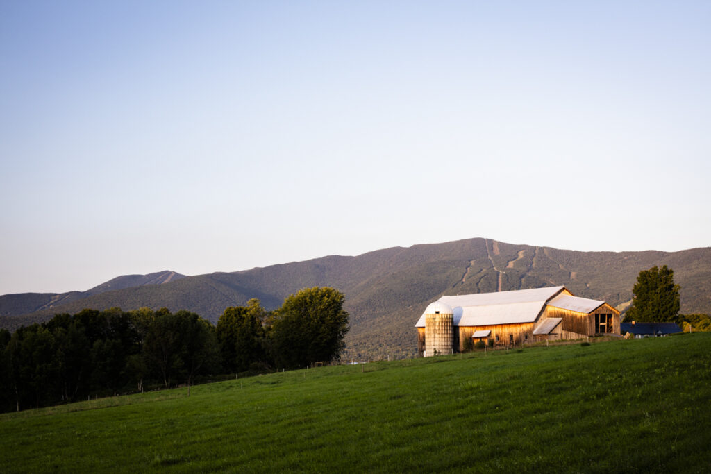 A scenic farm with a white-roofed barn and silo glows in the golden light of sunset, nestled against a backdrop of Vermont’s rolling mountains.