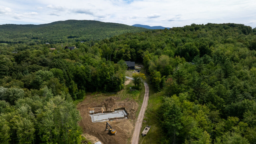 Aerial view of a home construction site with foundation work and an excavator, surrounded by dense forest and mountain scenery.