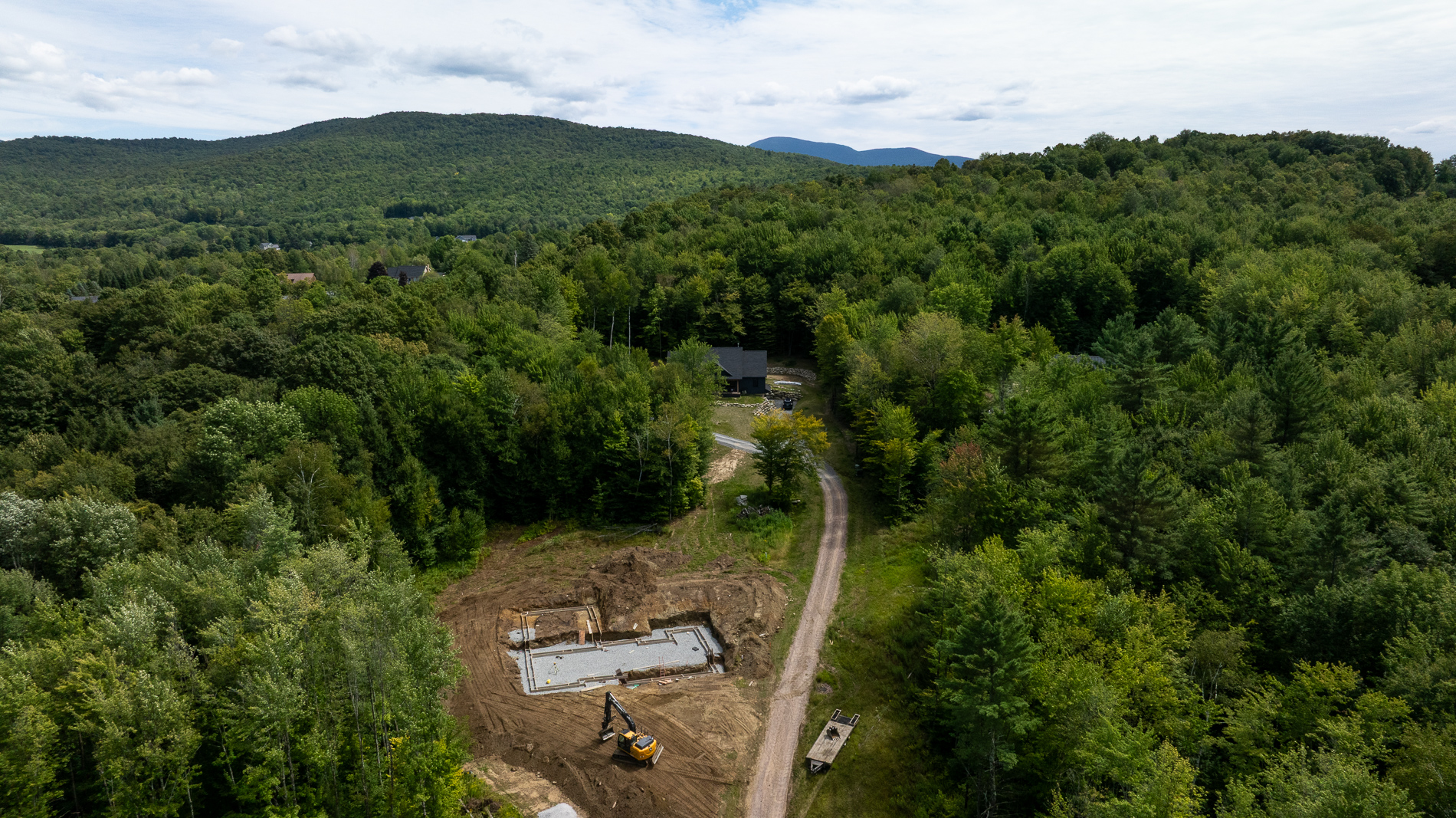 Aerial view of a home construction site with foundation work and an excavator, surrounded by dense forest and mountain scenery.