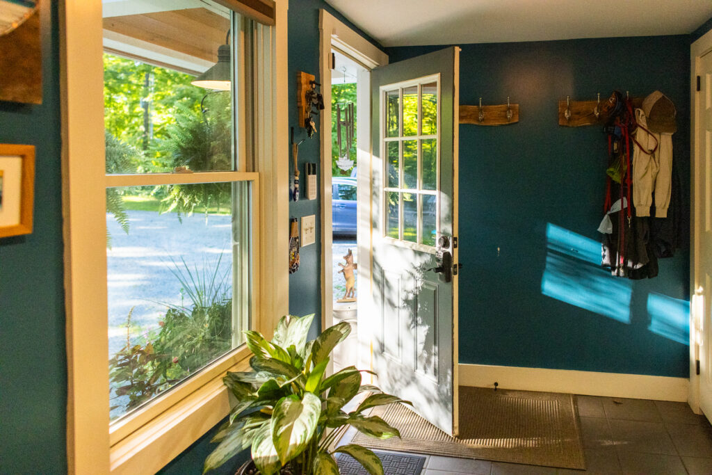 Sunlight streams into a cozy home entryway with a blue wall, coat hooks, houseplants, and an open front door leading outside.