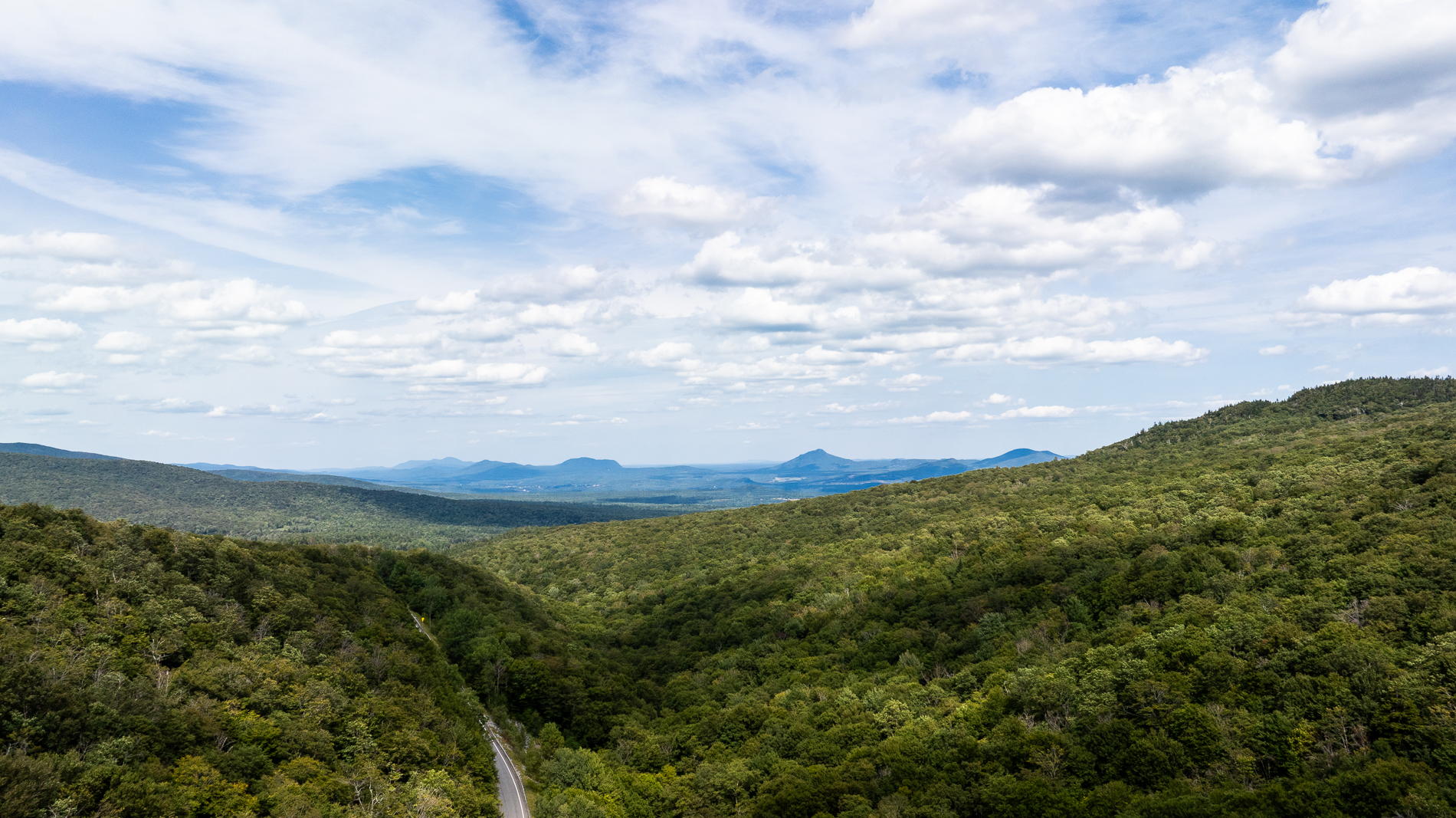 A sweeping view of Vermont’s lush Green Mountains under a partly cloudy sky, with a winding country road cutting through the dense forest.