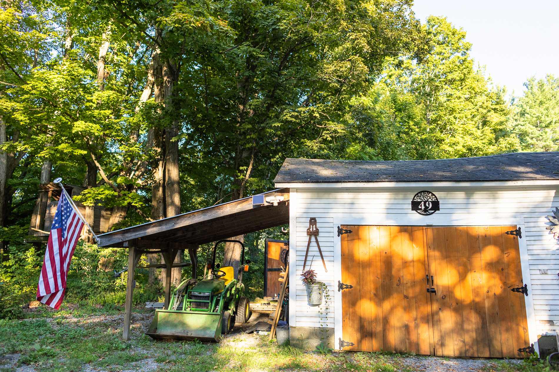 Green tractor parked beside a white shed with wooden doors and an American flag, surrounded by trees and morning sunlight.
