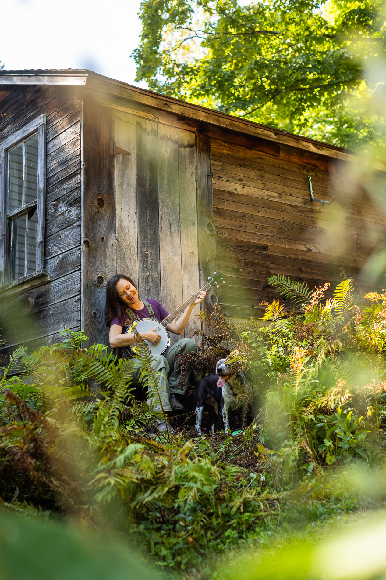 A woman sits on the steps of a rustic wooden cabin, smiling as she plays a banjo. Ferns surround the area, and a black-and-white dog stands nearby, looking content.