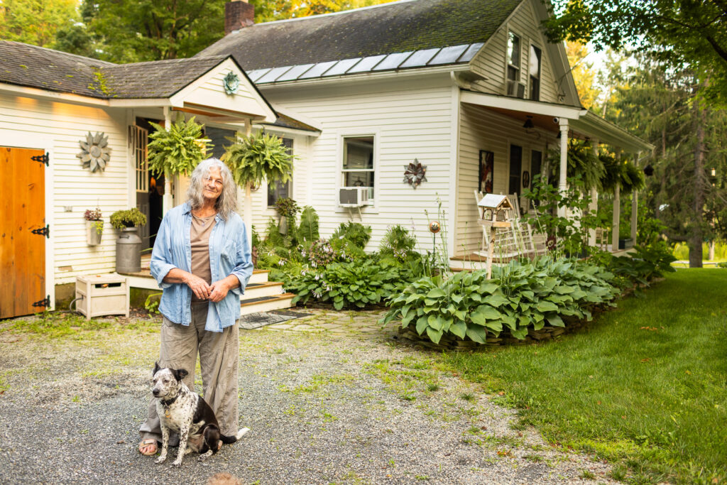 Older woman stands outside a white farmhouse-style home with her dog, surrounded by lush plants and a gravel walkway.