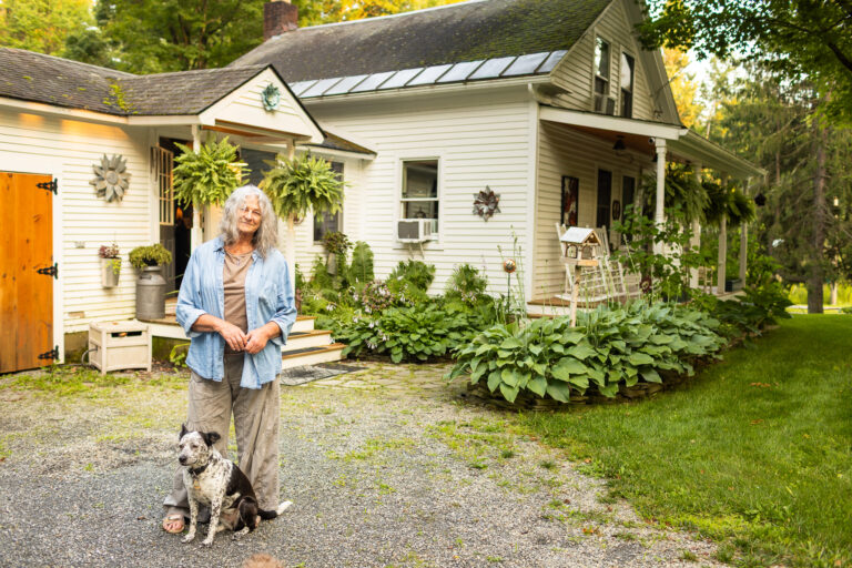 Older woman stands outside a white farmhouse-style home with her dog, surrounded by lush plants and a gravel walkway.