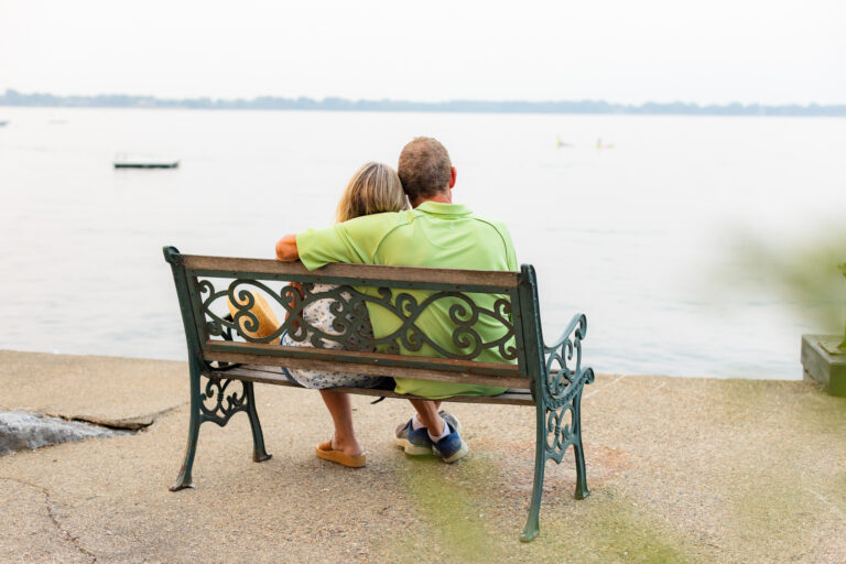 Older couple sits close together on a bench overlooking a calm lake, suggesting peace, connection, and a relaxed retirement.