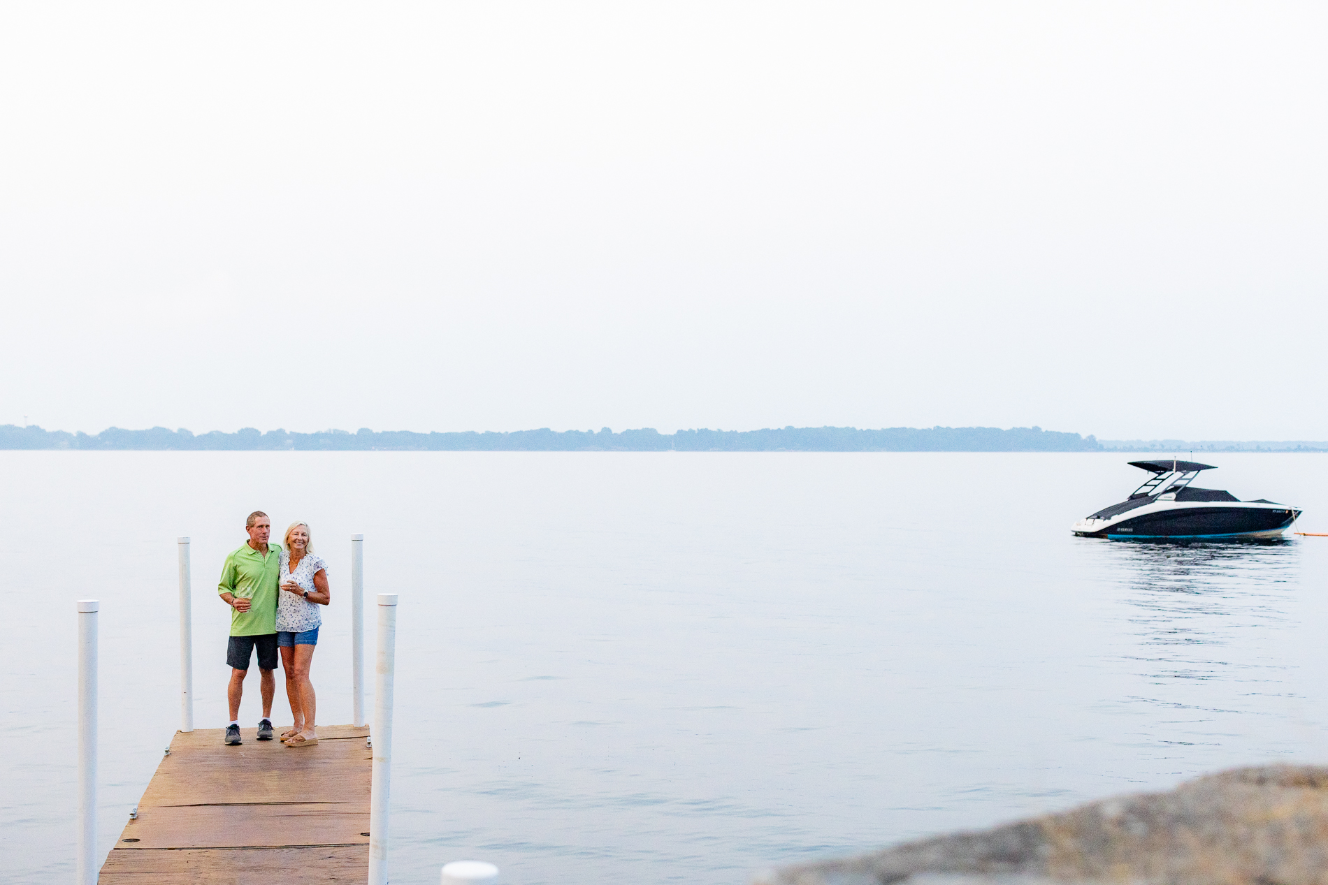 An older couple stands at the end of a dock, smiling and holding drinks, with a sleek black boat floating nearby on a calm lake under a pale sky.