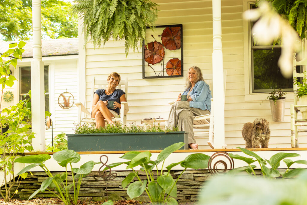 Two older women relax with coffee on a front porch surrounded by greenery, with a small dog nearby and potted plants in view.