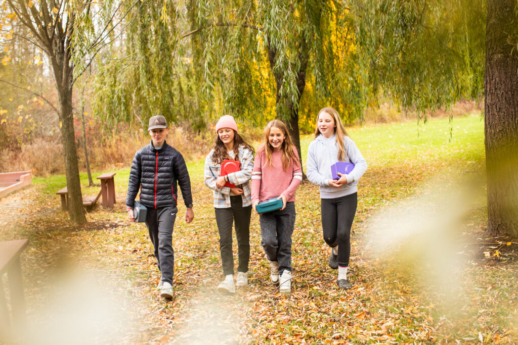 Four smiling kids walk together through a park in autumn, carrying colorful boxes and bundled in casual, cool-weather clothing.