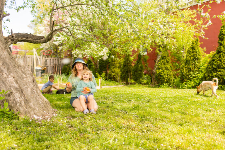 Smiling parent sits under a blooming tree with young children and a dog nearby in a sunny backyard garden, holding fresh-picked fruit.