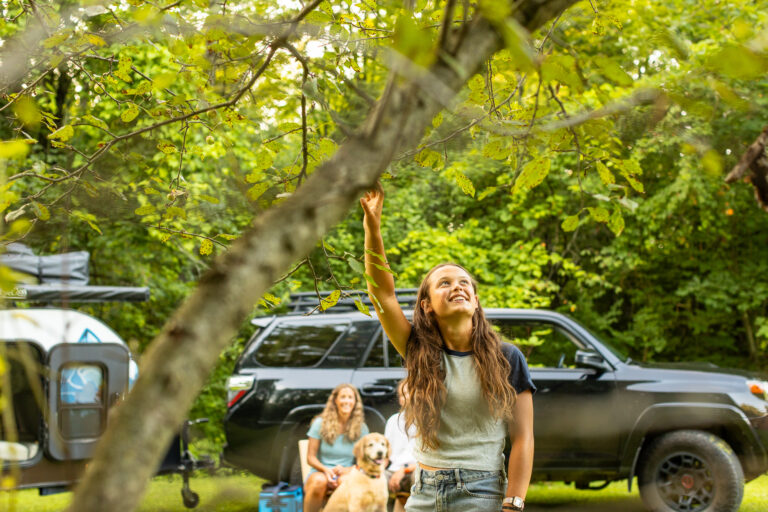 Smiling girl reaches for a tree branch at a campsite, while her family, dog, SUV, and camper are visible in the background.