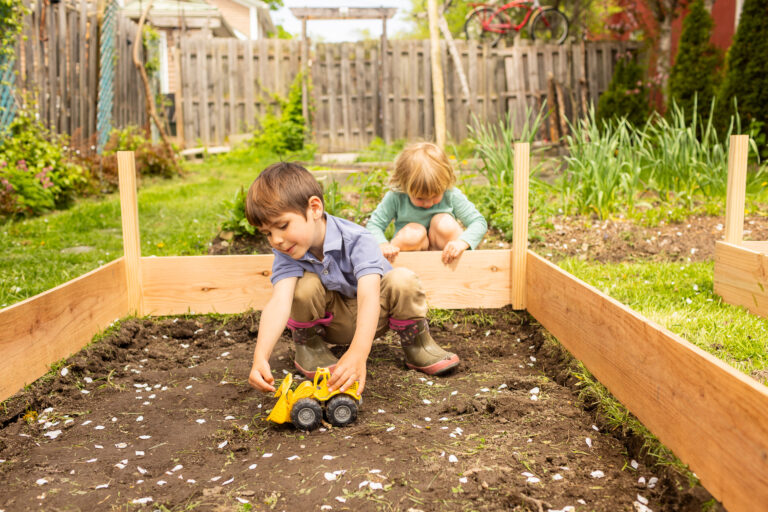 Two young children play in a backyard garden bed; one uses a toy bulldozer in the soil while the other explores behind him. The scene is bright and natural, with wood-raised beds and a fence in the background.