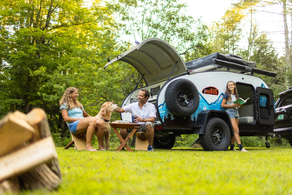 Family camping scene with a small trailer in a grassy, wooded area. A man and woman sit at a picnic table with a golden retriever, smiling and enjoying the outdoors. A teenage girl stands by the camper holding a book and smiling.