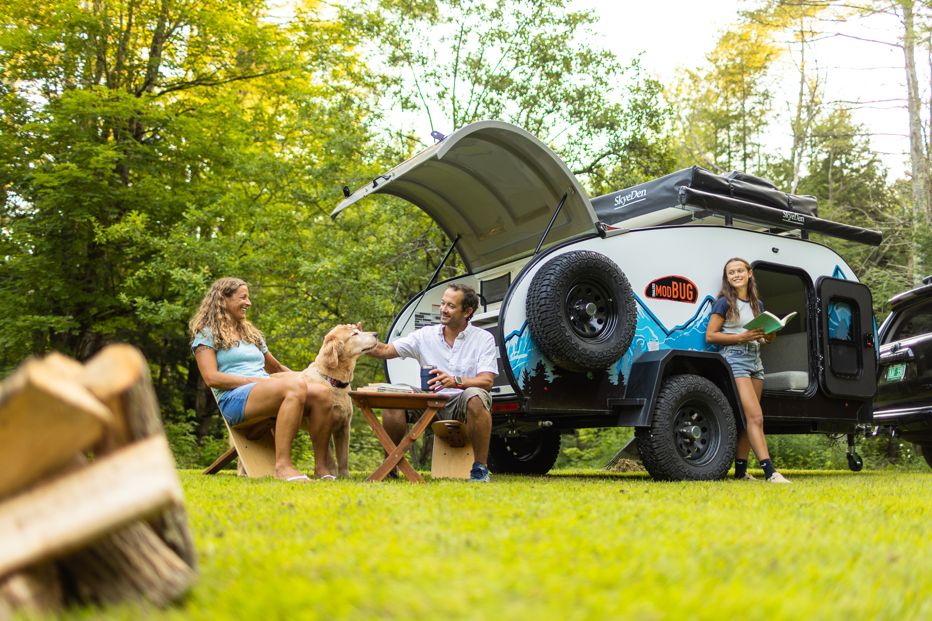 Family camping scene with a small trailer in a grassy, wooded area. A man and woman sit at a picnic table with a golden retriever, smiling and enjoying the outdoors. A teenage girl stands by the camper holding a book and smiling.