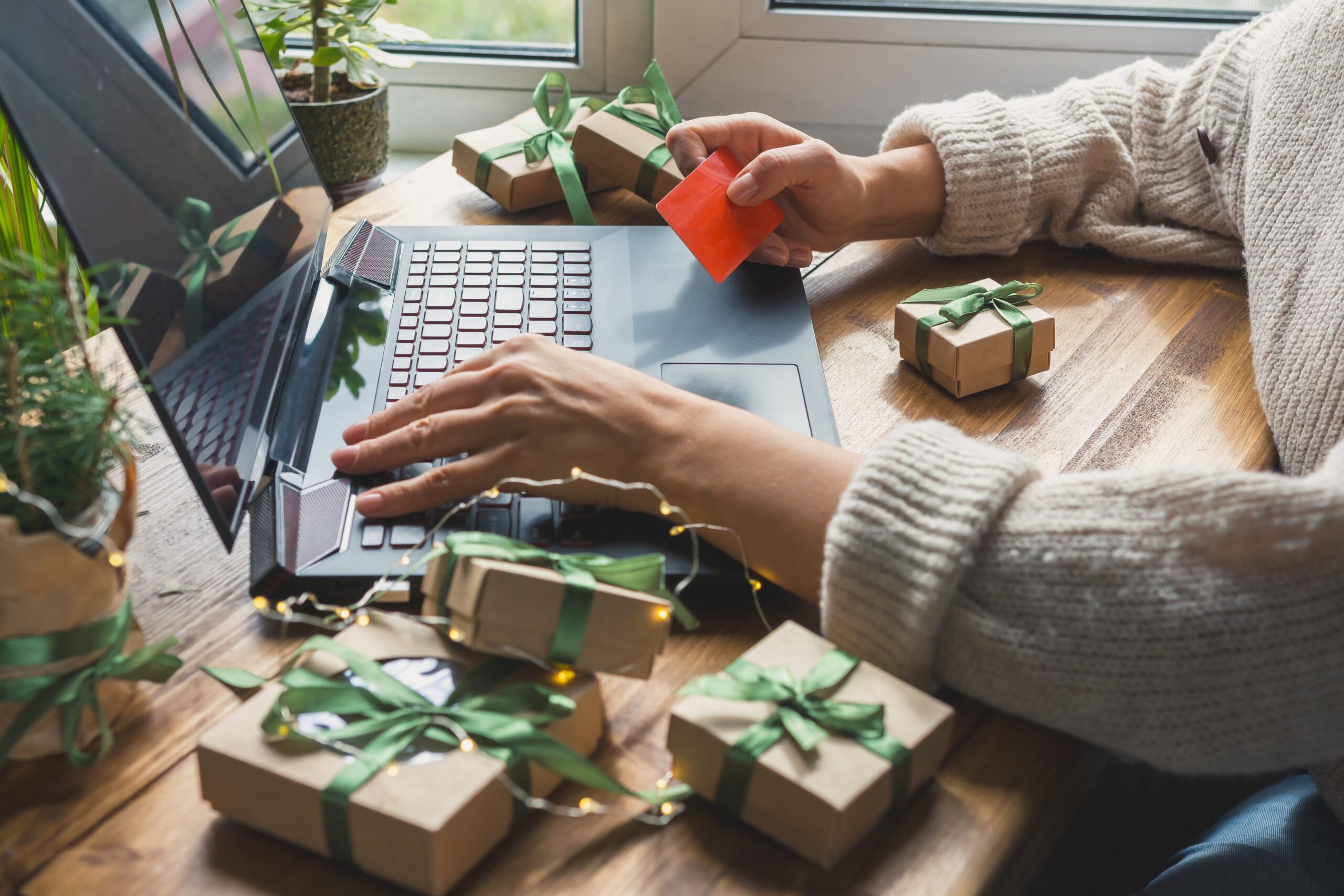 At a small table cluttered with tiny gift boxes adorned with green ribbon bows, a woman in a thick beige sweater looks at a red payment card in her hand and types on an open black laptop.