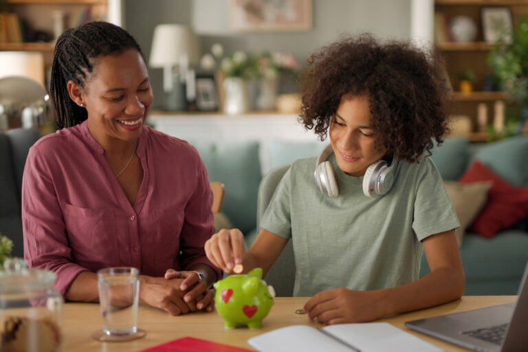 Image Alt Text A mother and her child are sitting at a table, smiling as they deposit coins into a green piggy bank. The child, wearing headphones around their neck, is placing a coin into the piggy bank while the mother watches encouragingly.