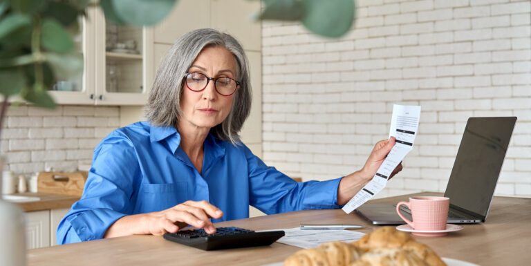 In a relaxing cream-colored kitchen, a woman with silver hair, glasses, and a bright blue shirt is typing on a calculator while reviewing a financial statement, with a laptop, coffee cup, and pastries on the table in front of her.